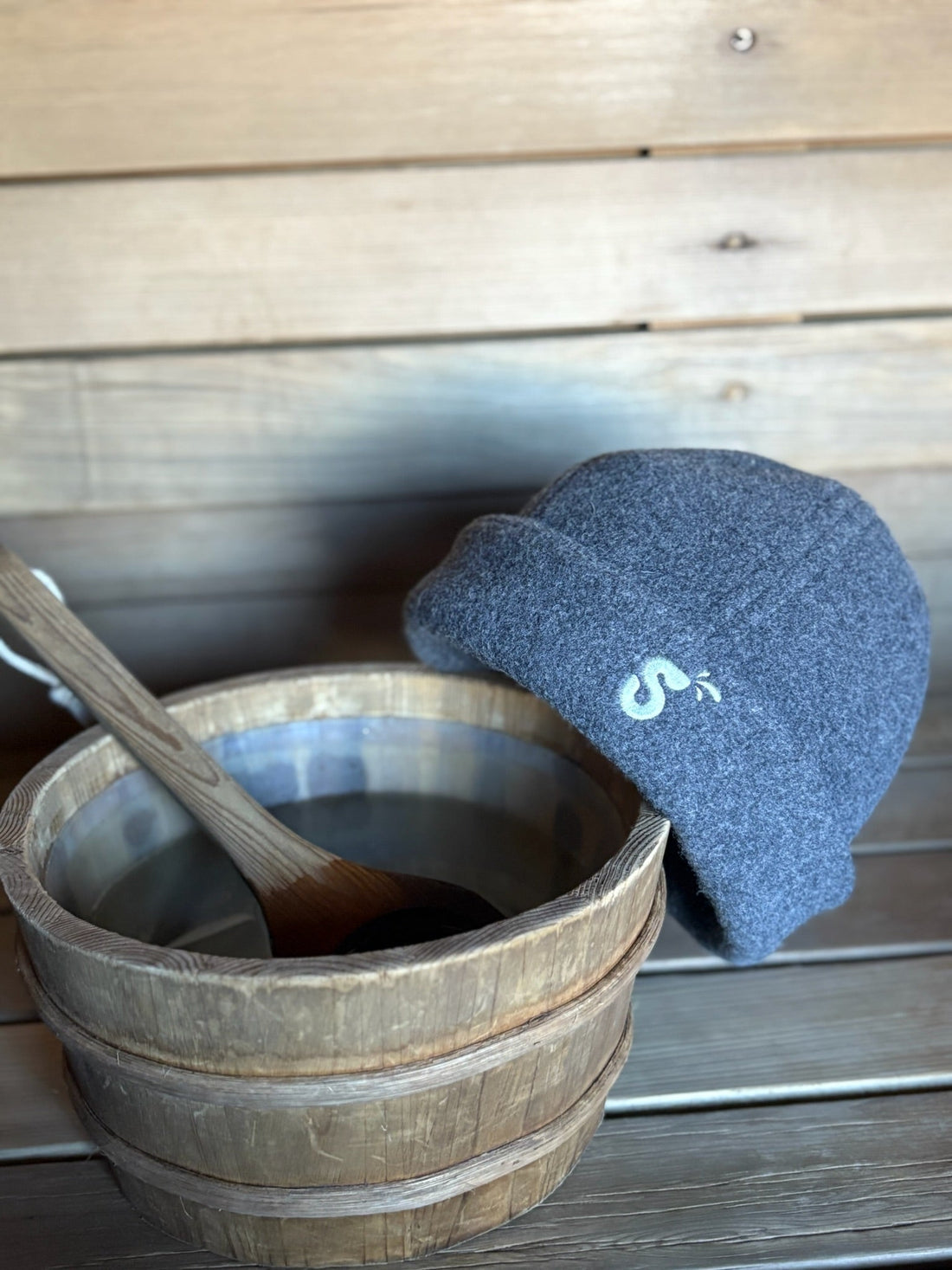 Charcoal gray Schvitzin wool felt sauna hat leaning against a wooden sauna bucket and ladle on a cedar bench