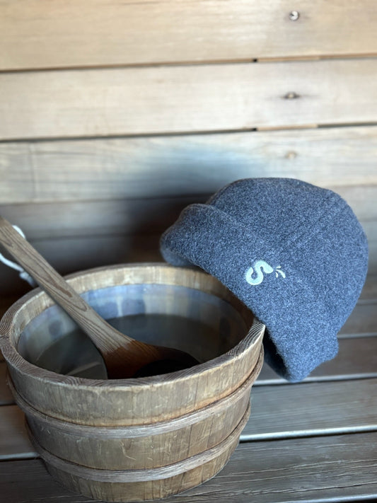 Charcoal gray Schvitzin wool felt sauna hat leaning against a wooden sauna bucket and ladle on a cedar bench