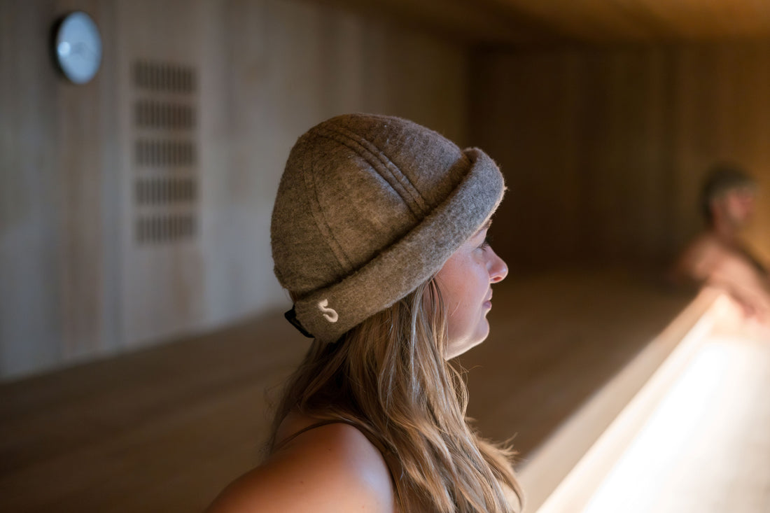 Woman wearing a brown merino wool sauna hat inside a wood-paneled sauna, profile view, warm ambient light