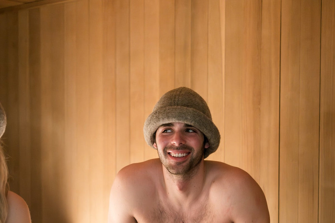 Smiling man wearing a charcoal gray Schvitzin merino wool felt sauna hat on a cedar bench inside a wood-paneled sauna