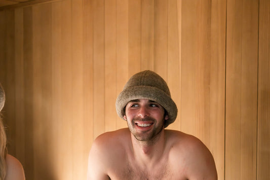 Smiling man wearing a charcoal gray Schvitzin merino wool felt sauna hat on a cedar bench inside a wood-paneled sauna