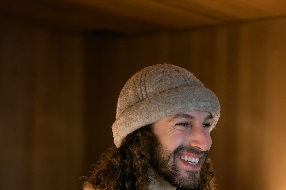 A bearded man with long curly hair smiles warmly while wearing a charcoal gray Merino wool Schvitzin sauna hat inside a wood-paneled sauna, lit with a warm amber glow