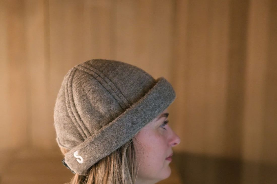 Side profile of a woman wearing a charcoal gray Schvitzin merino wool felt sauna hat with embroidered "S" logo inside a wood-paneled sauna