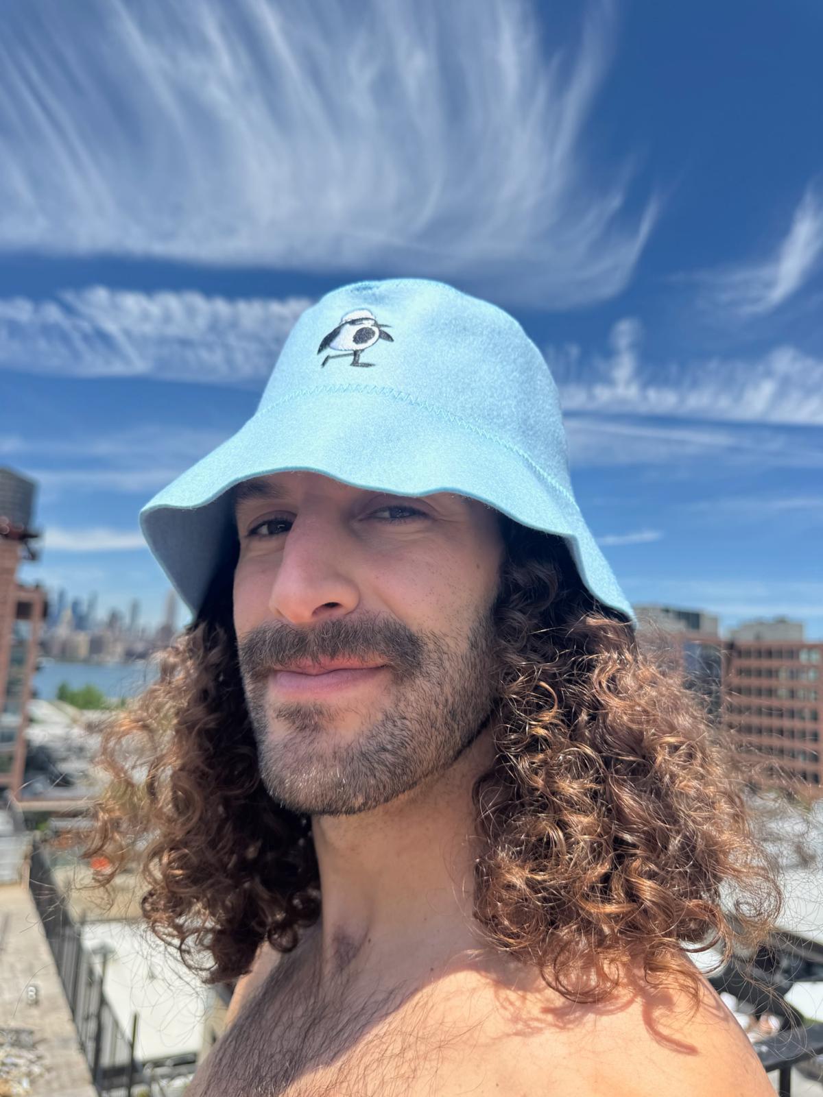  Person wearing the Sky colorway Schvitzin Starter merino wool sauna hat on a Brooklyn rooftop with the Manhattan skyline in the background