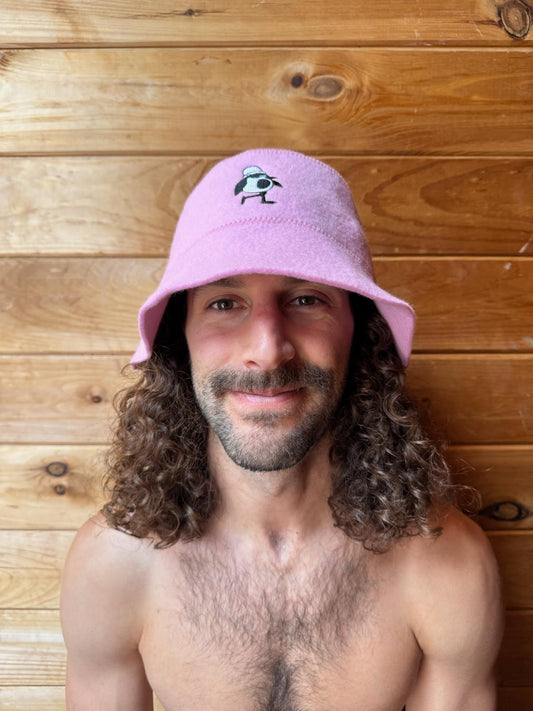 Person wearing the Rose colorway Schvitzin Starter merino wool sauna hat inside a sauna with wood panel background