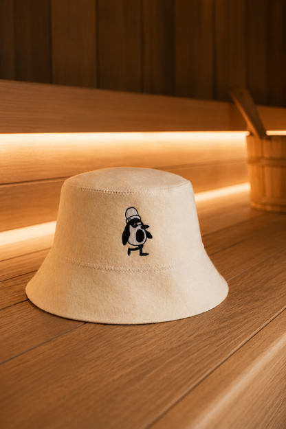 Cream merino wool sauna hat with an embroidered penguin resting on a wooden sauna bench, a wooden bucket beside it.