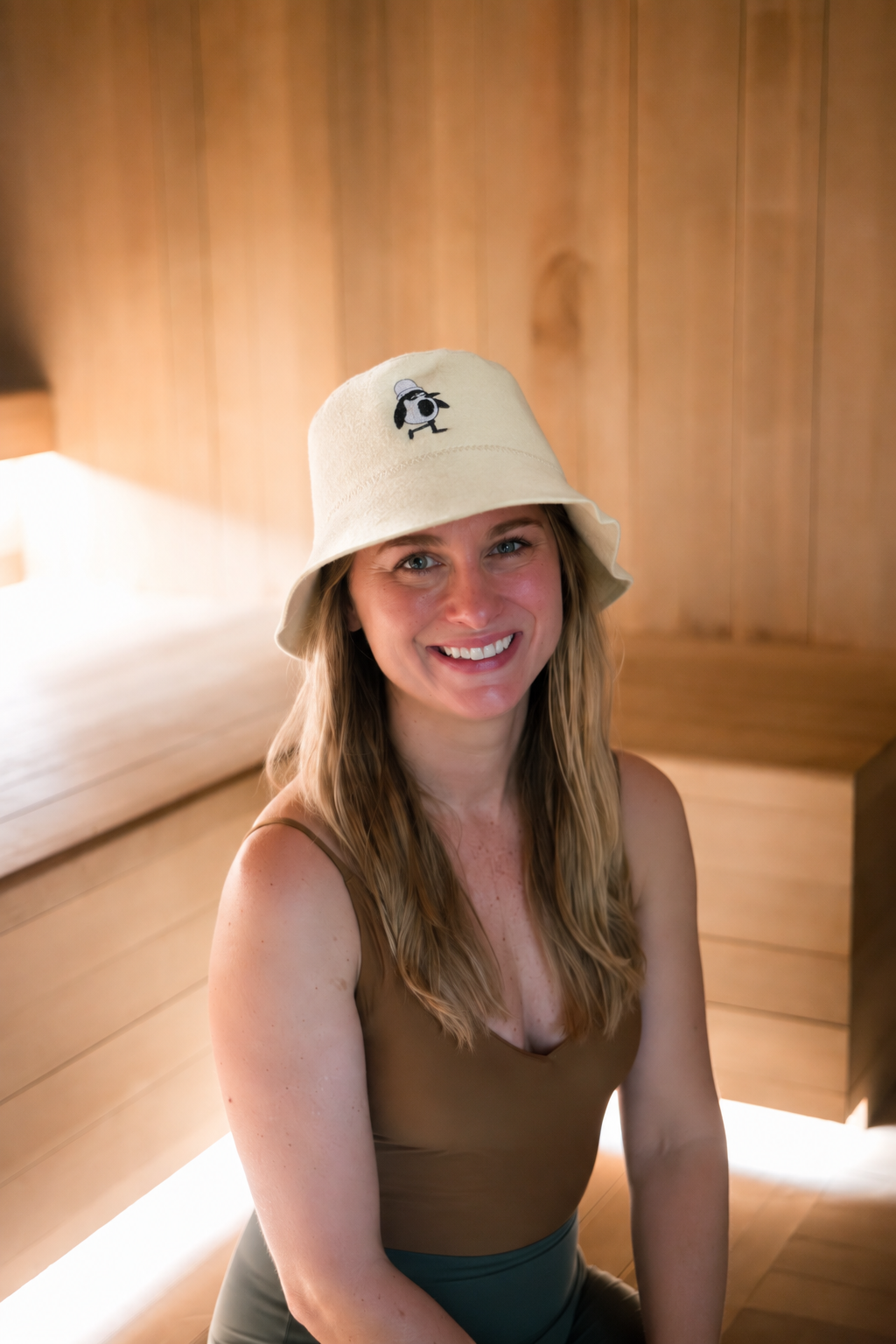 Smiling woman wearing a cream merino wool sauna hat with embroidered penguin, seated in a wood-paneled sauna.