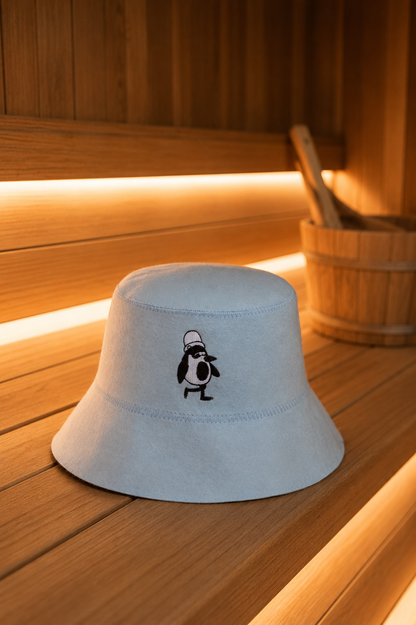Light blue merino wool sauna hat with an embroidered penguin on a wooden sauna bench, a wooden bucket beside it.