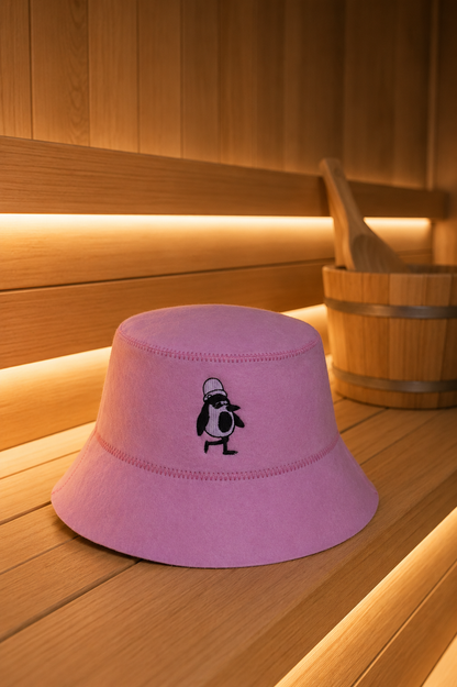 Rose merino wool sauna hat with an embroidered penguin on a wooden sauna bench, a wooden bucket beside it.