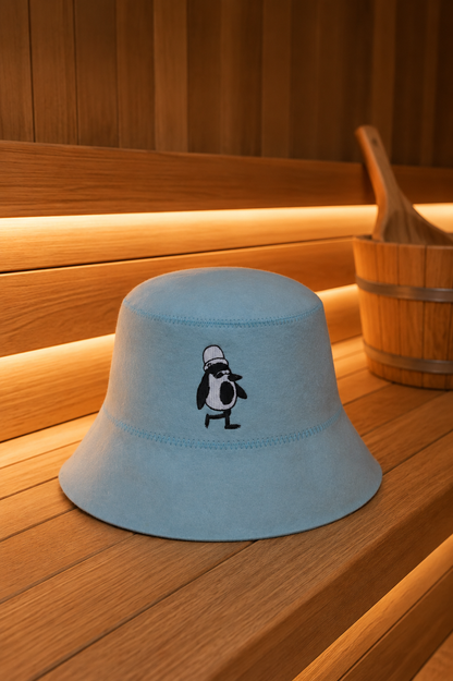 Steam blue merino wool sauna hat with an embroidered penguin on a wooden sauna bench, a wooden bucket beside it.