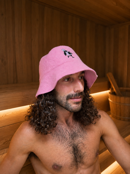 Man with long curly hair wearing a pink merino wool sauna hat with embroidered penguin, seated in a wood-paneled sauna.