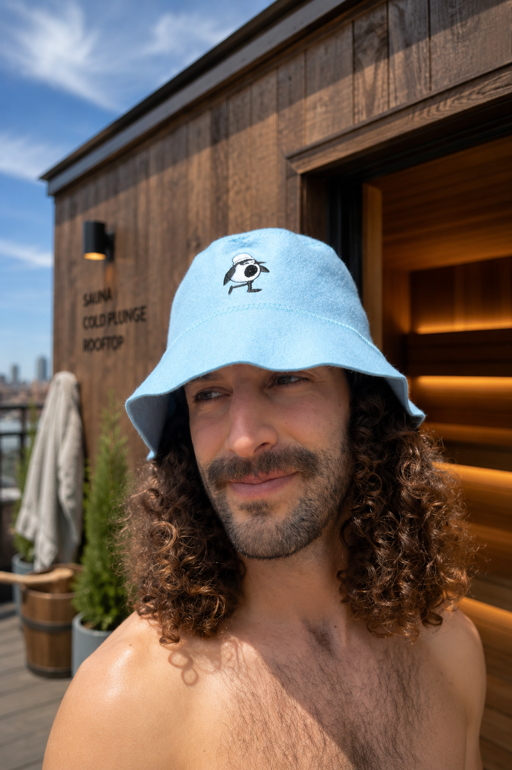 Man with curly hair wearing a light blue merino wool sauna hat with embroidered penguin on a rooftop sauna deck.